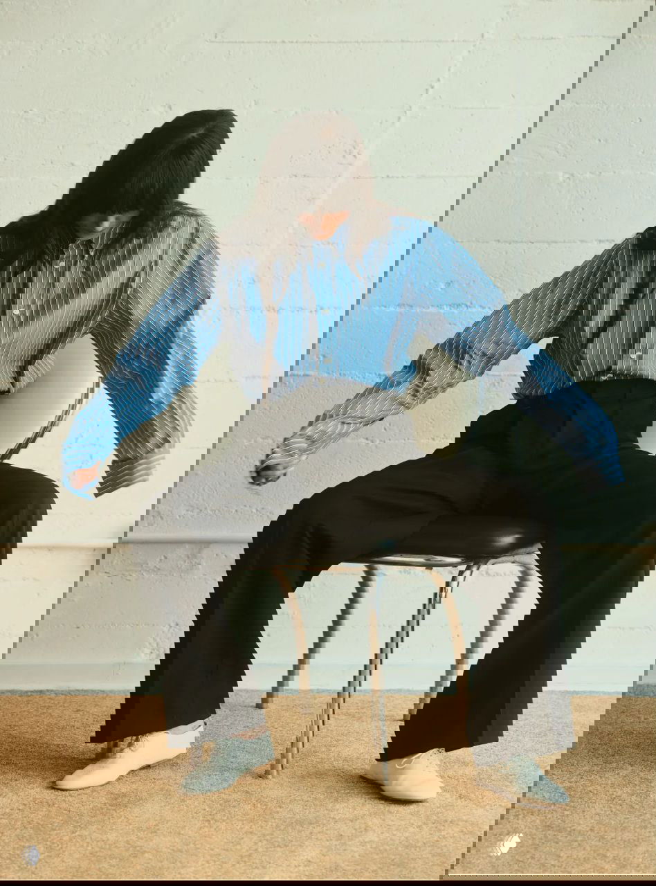 Актриса сидит в рубашке в полоску и расклешённых брюках; кадр подчёркивает расслабленную позу и акцент на силуэте одежды.