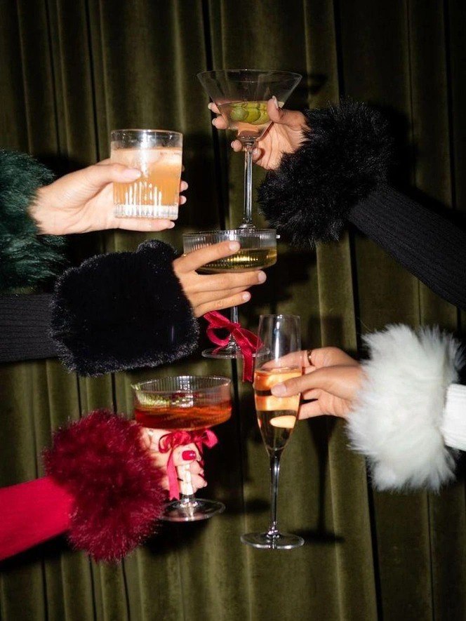 Руки в тёплых меховых манжетах поднимают бокалы с коктейлями на фоне зелёной бархатной драпировки — атмосфера праздничного тоста.