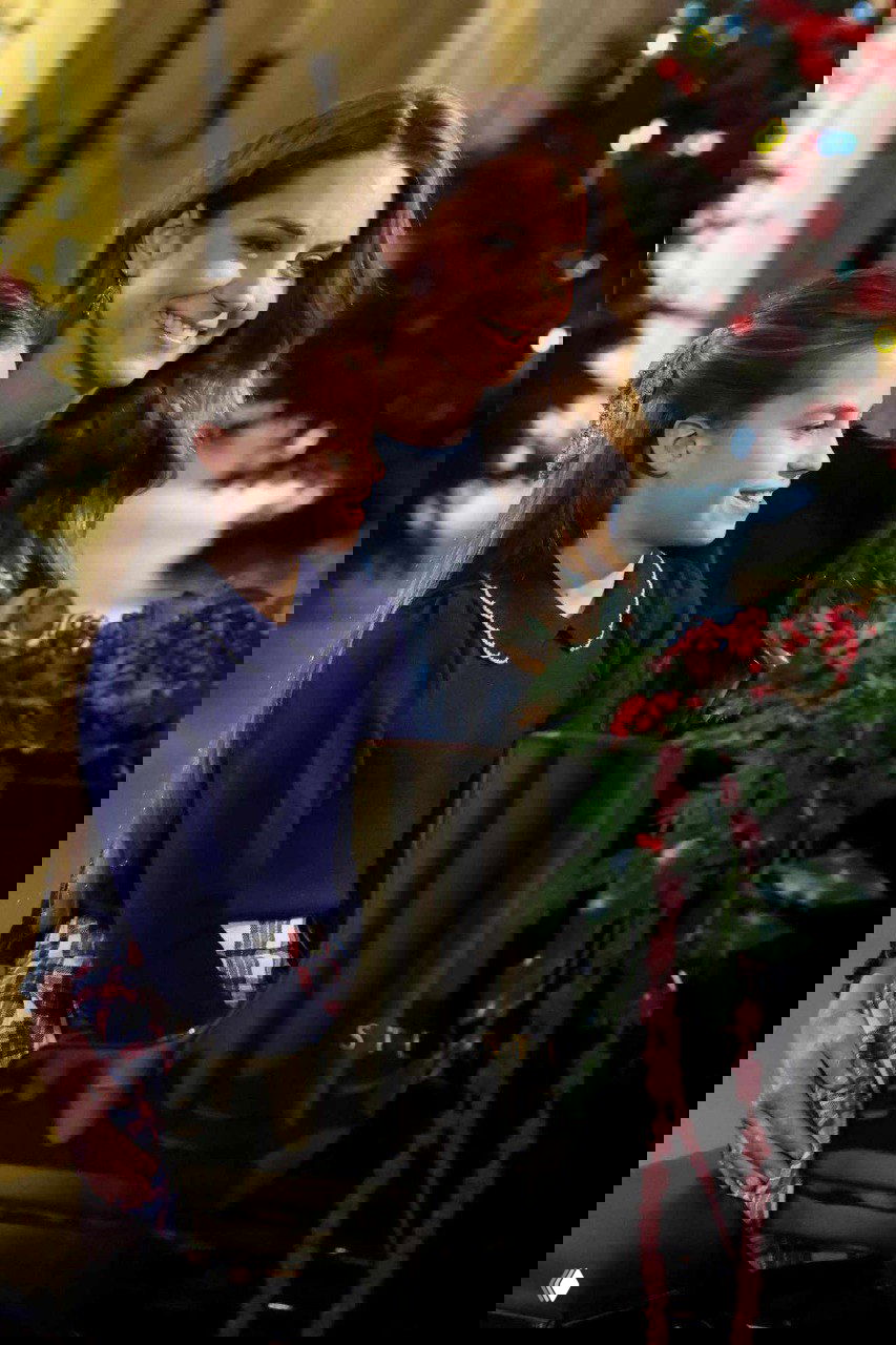 Женщина и девочка улыбаются у рояля в праздничной атмосфере; видны ёлочные декорации, тёплое освещение и праздничные детали интерьера зала.