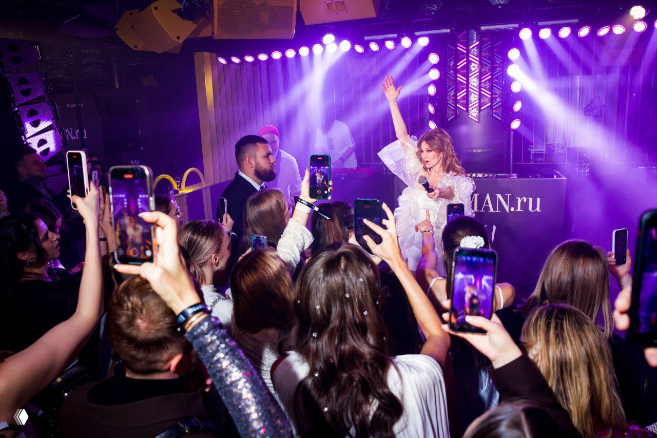 Зрители с поднятыми смартфонами снимают выступление певицы на сцене с пурпурными и белыми световыми эффектами в клубной атмосфере.