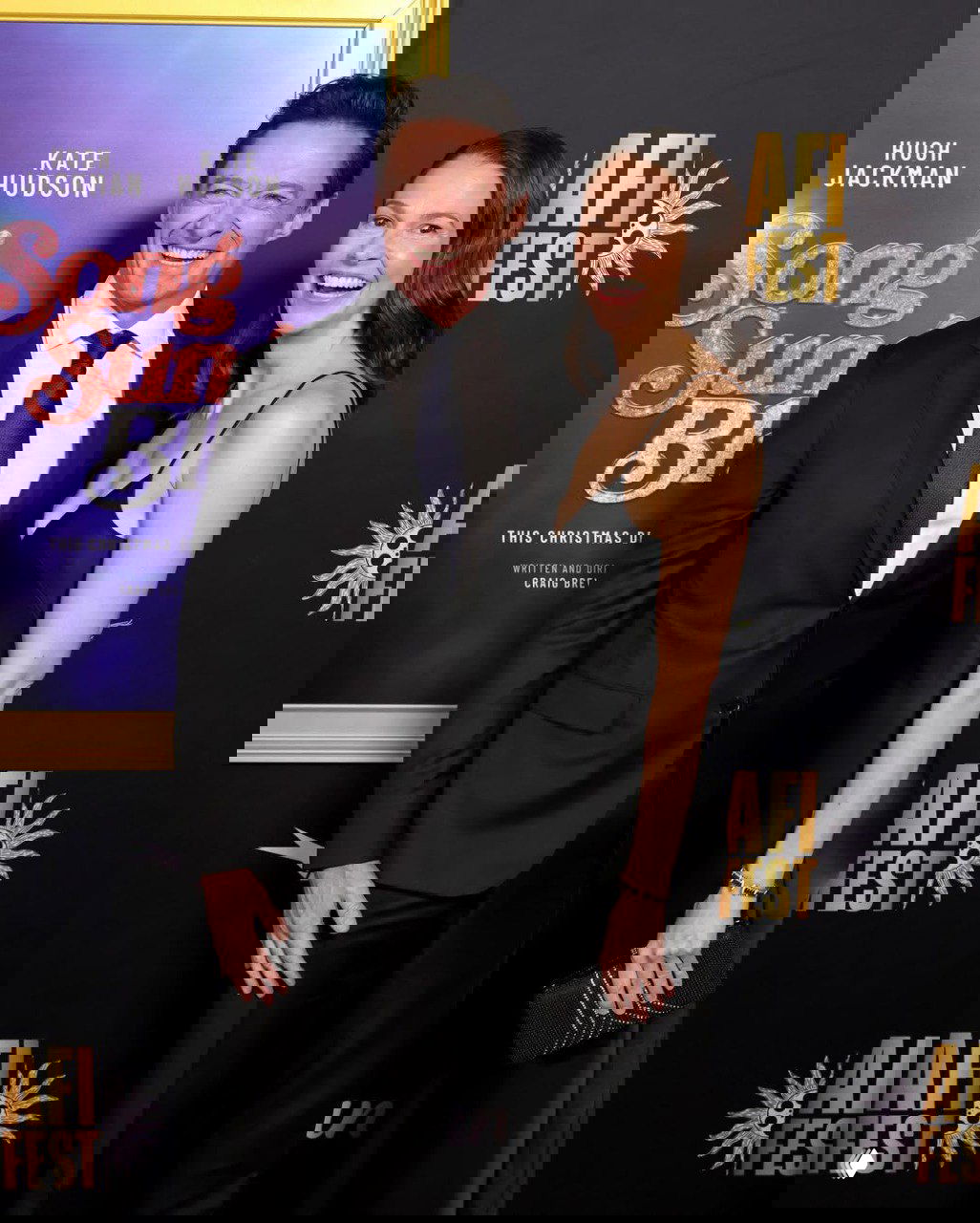 Мужчина и женщина в вечерних нарядах улыбаются на красной дорожке у плаката премьеры фильма «Песня звучит печально» в Лос‑Анджелесе.