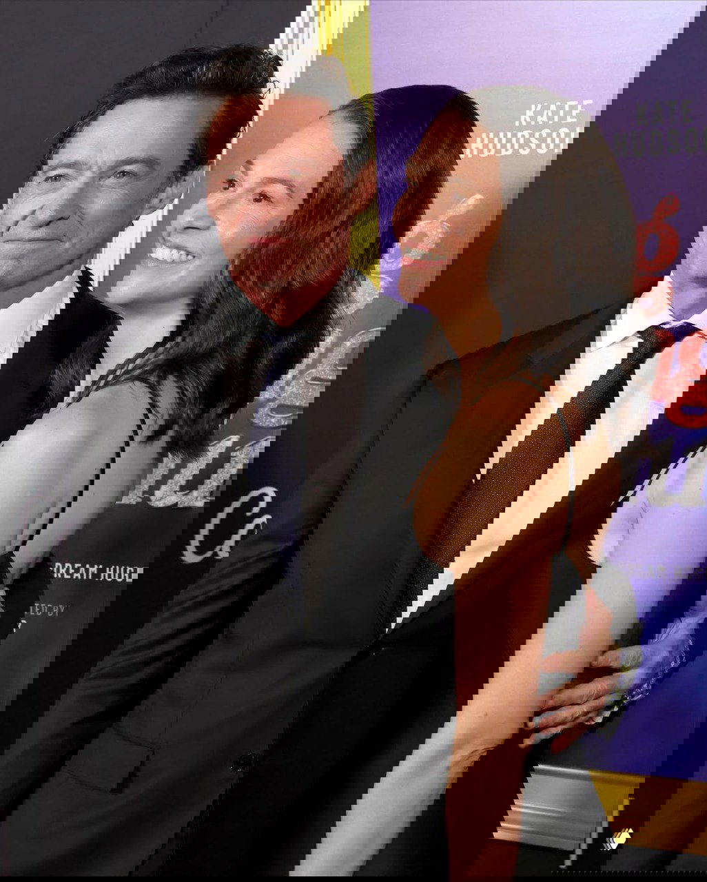 Крупный план пары на красной дорожке: мужчина в тёмном костюме и женщина в чёрном платье улыбаются на фоне постера премьеры в Лос‑Анджелесе.