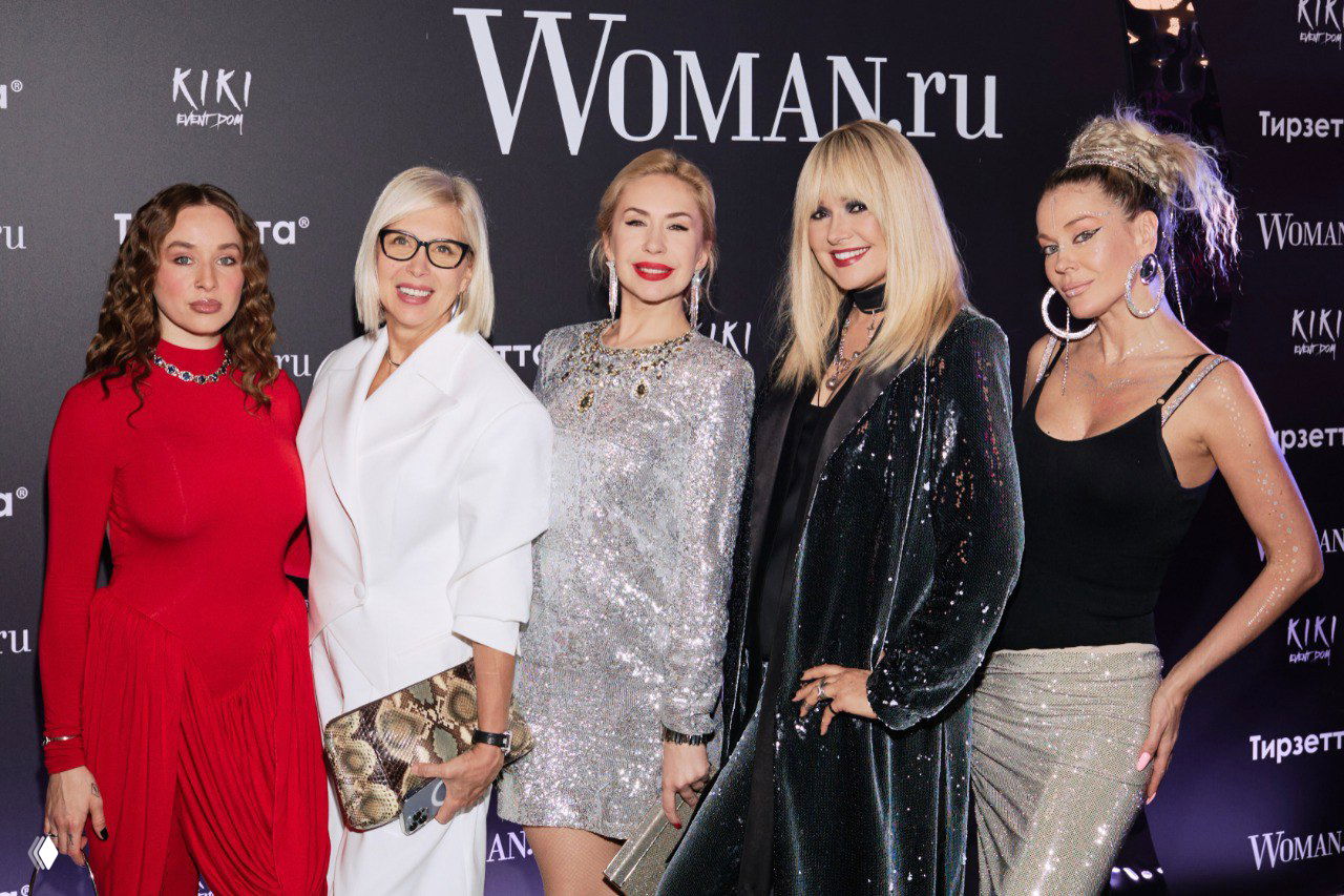 Группа знаменитостей и гостей на красной дорожке перед баннером Woman.ru: несколько женщин в вечерних нарядах и блёстках.