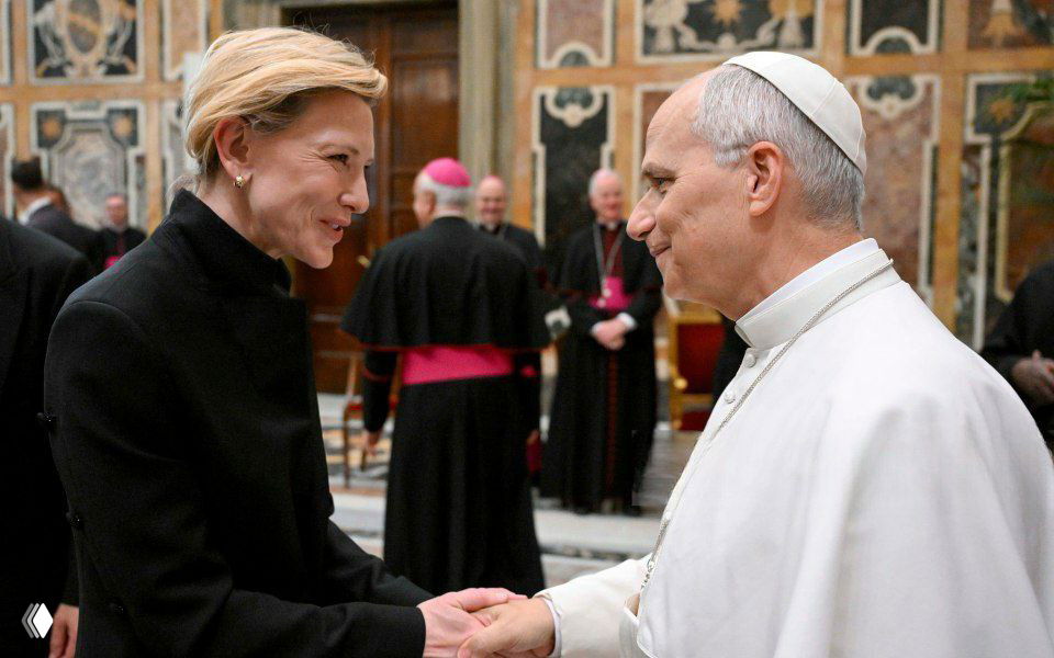 Кейт Бланшетт пожимает руку Папе Римскому в зале Ватикана; кадр показывает близкий обмен приветствиями на официальной церемонии.