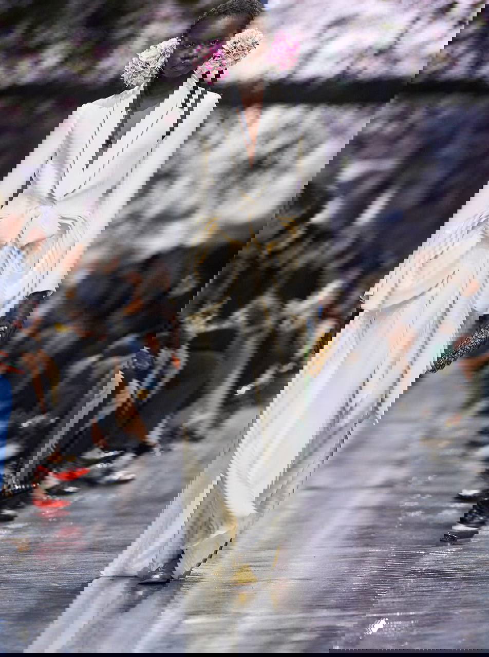 Белое платье с перьями и сложным драпированием; модель на показе первой кутюрной коллекции Джонатана Андерсона для Dior, цветочный фон подиума.