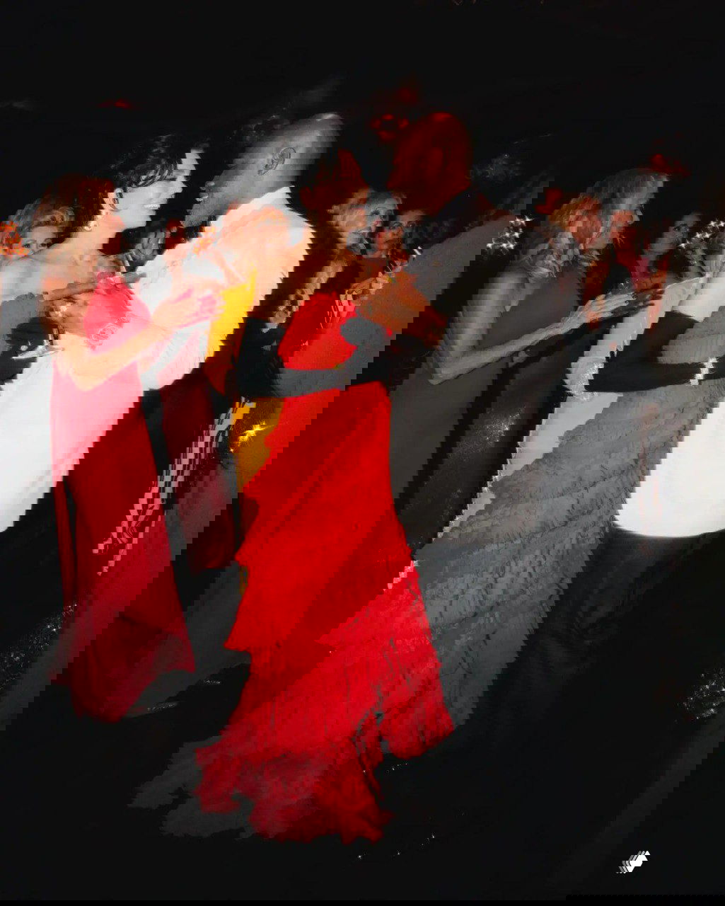 Пара и гости на танцполе: женщина в многоярусном красном платье в обнимку с мужчиной в светлом пиджаке, вокруг люди в вечерних нарядах.