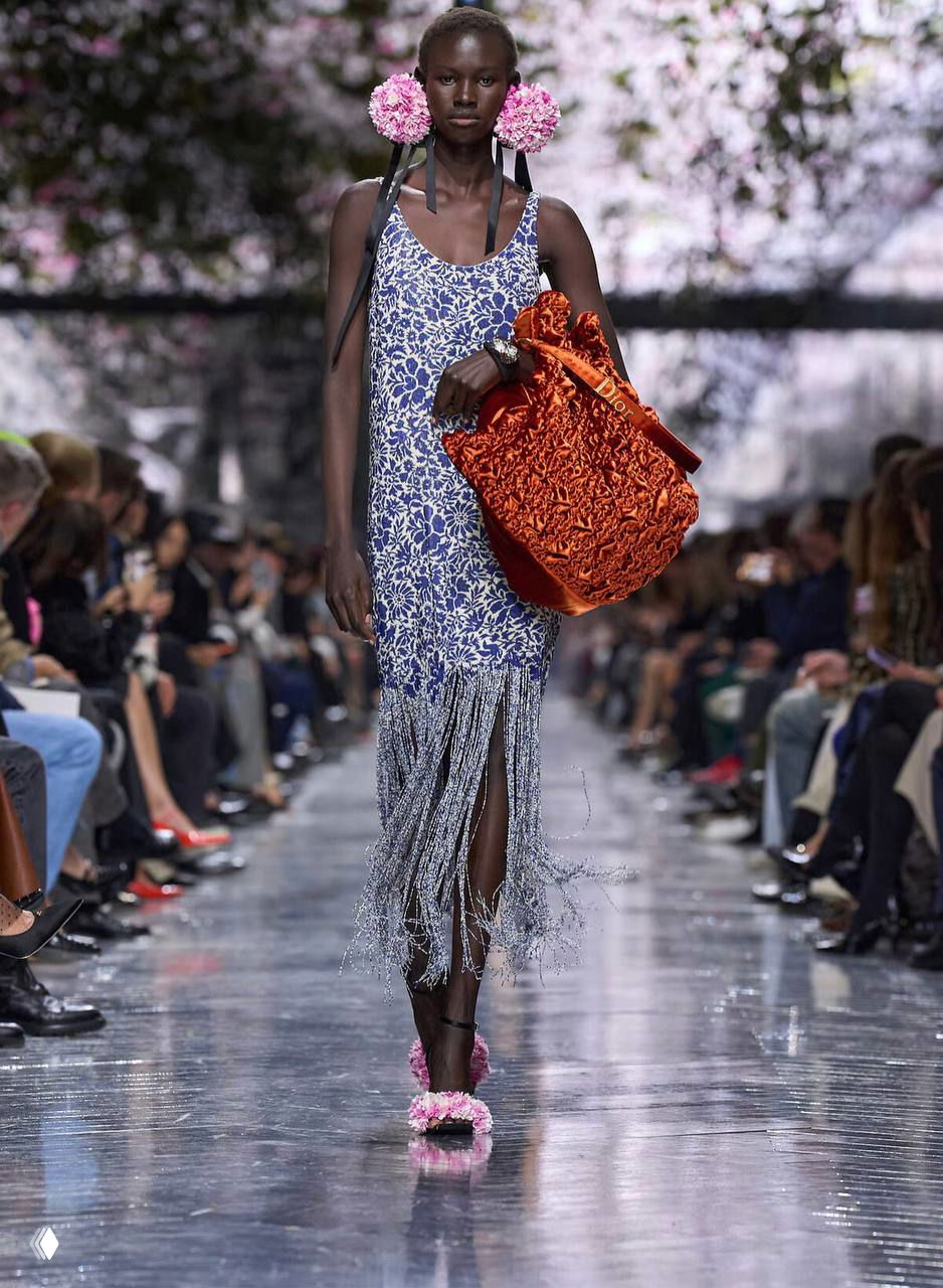 Лёгкое платье с бахромой и крупной оранжевой сумкой; модель на шоу Dior Джонатана Андерсона, заметны цветочные аксессуары в прическе.