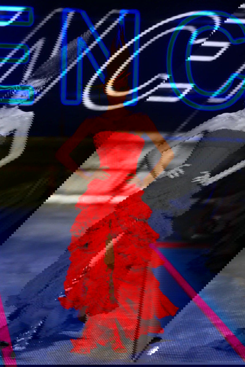 Подиумное изображение длинного красного платья с многоярусной юбкой на модели, на фоне крупного светящегося логотипа бренда.