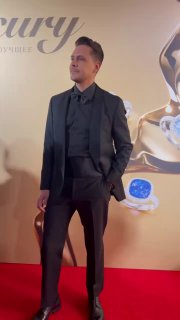 Мужчина в тёмном костюме и рубашке позирует на красной дорожке премии, на фоне — баннер и декор с ювелирными элементами.