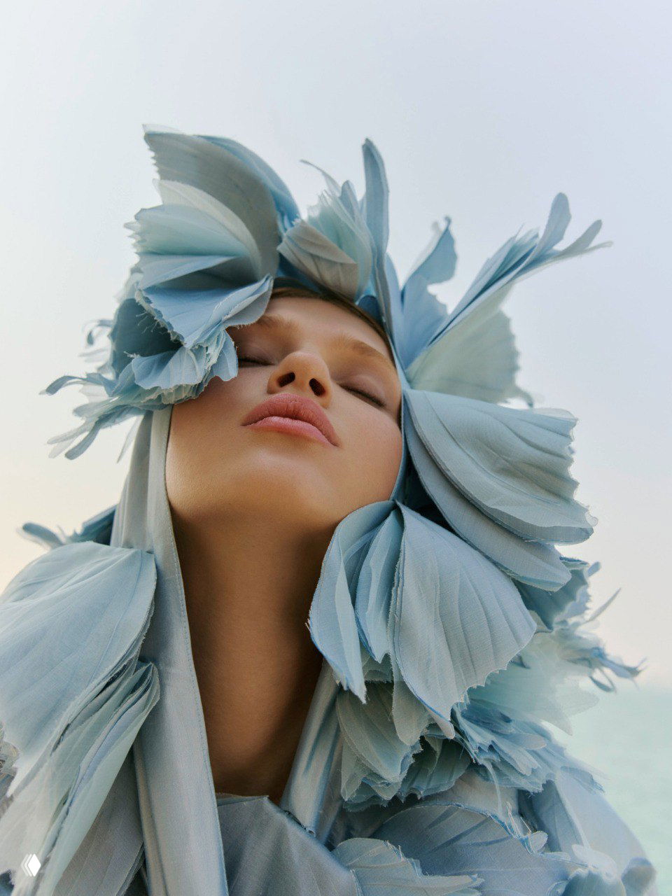 Крупный план модели в небесно-голубом текстильном головном уборе в форме лепестков — редакционная фэшн-съёмка с акцентом на образ и макияж.