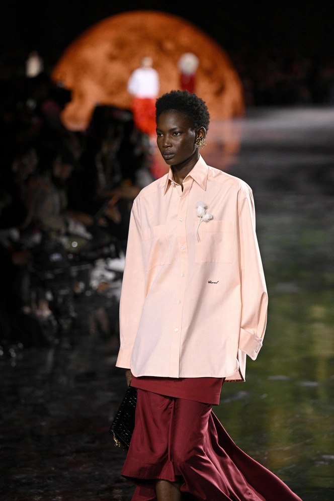 Темнокожая модель в свободной розовой рубашке и бордовой юбке с небольшой брошей у кармана на фоне сценографии показа.