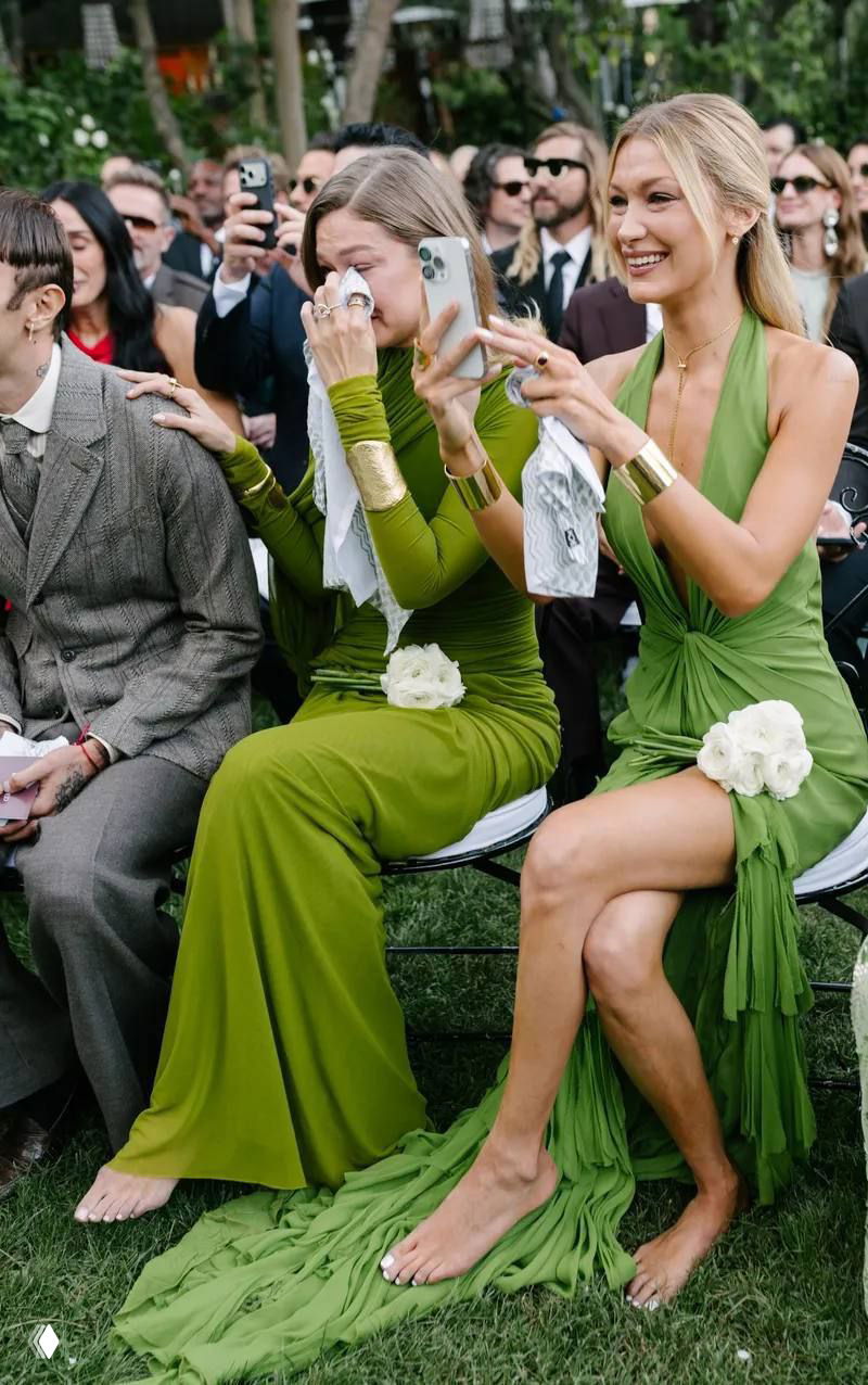 Гости в зелёных платьях на свадьбе: одна женщина плачет, другая улыбается и снимает событие на телефон в окружении гостей на открытой церемонии.