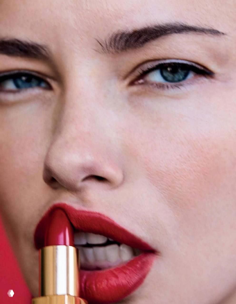 Крупный план: губы с ярко‑красной помадой и трубочка помады у губ на фоне лица модели — акцент на макияже и текстуре кожи.