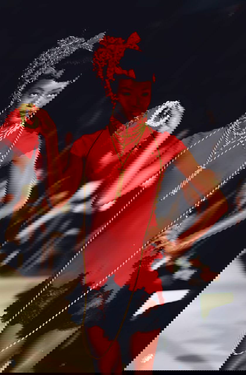 Темнокожая модель в красном платье с длинными золотыми цепями, клатчем и сценическим макияжем на показе Dior осень‑зима 1997, подиумный кадр.