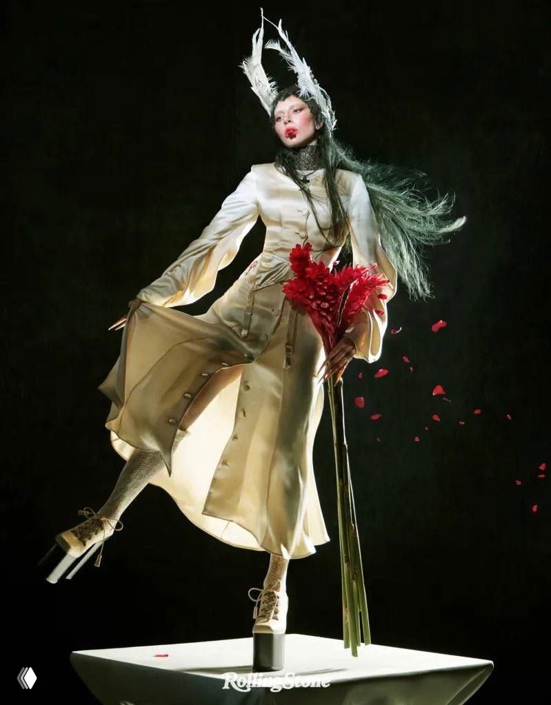 Студийный портрет женщины в авангардном белом платье и высоких платформах, держит пучок красных цветов на тёмном фоне.