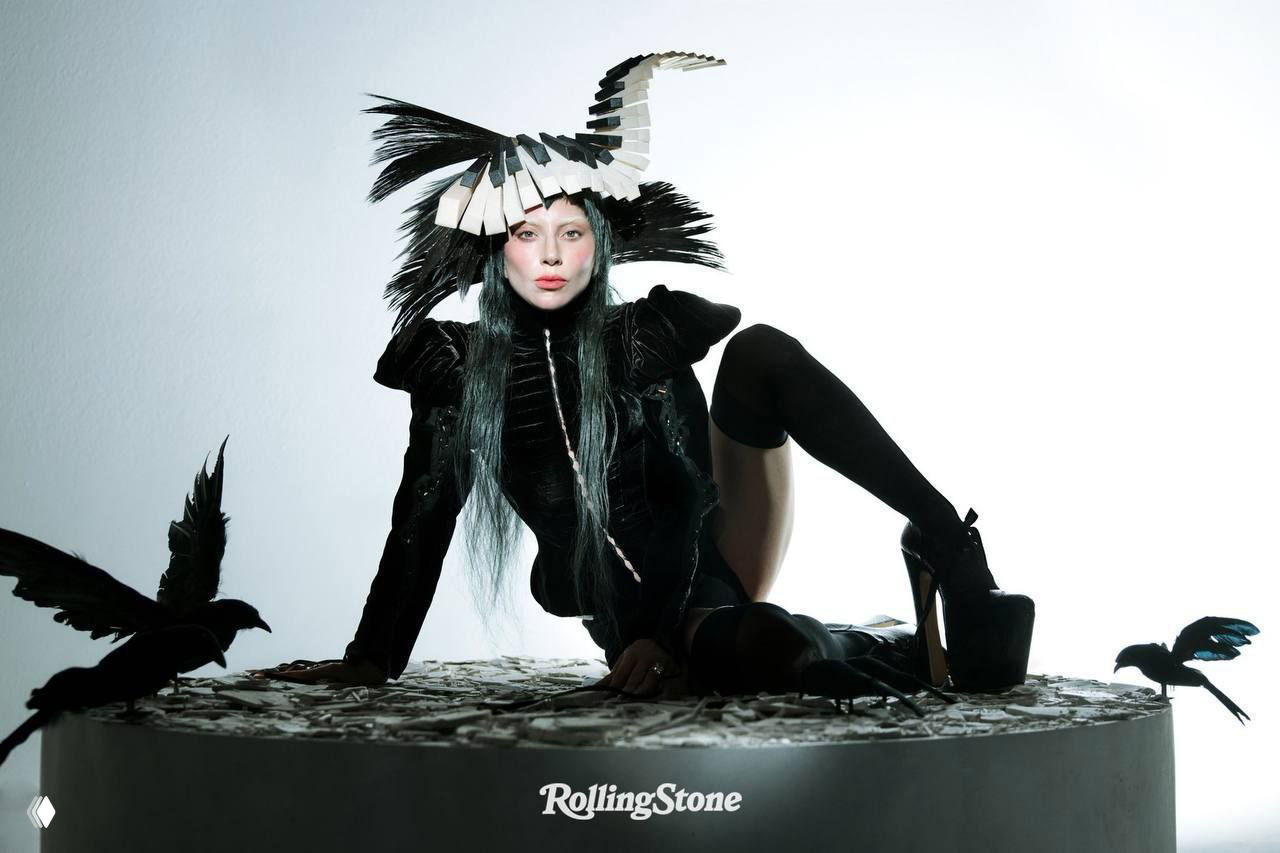 Женщина в чёрном костюме с головным убором и декорациями в виде птиц на светлом фоне, постановочный fashion-кадр.