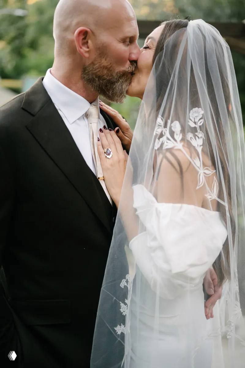 Крупный план поцелуя: жених целует невесту под вуалью с цветочной вышивкой — интимный кадр со свадебной церемонии на свежем воздухе.