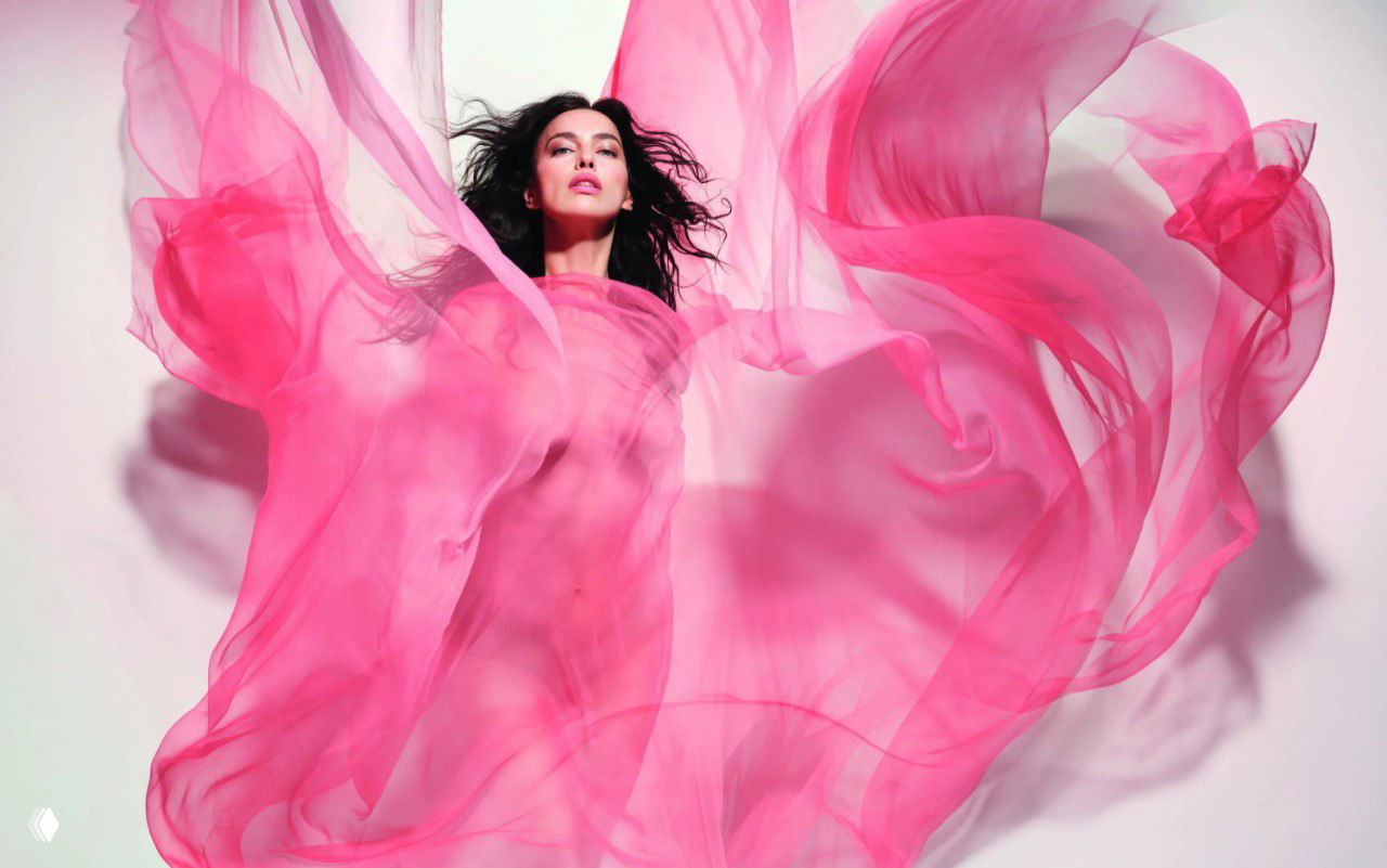 Модель в розовом струящемся наряде, ткань развевается вокруг тела, кадр передаёт лёгкость и ассоциации с цветами и ветром.
