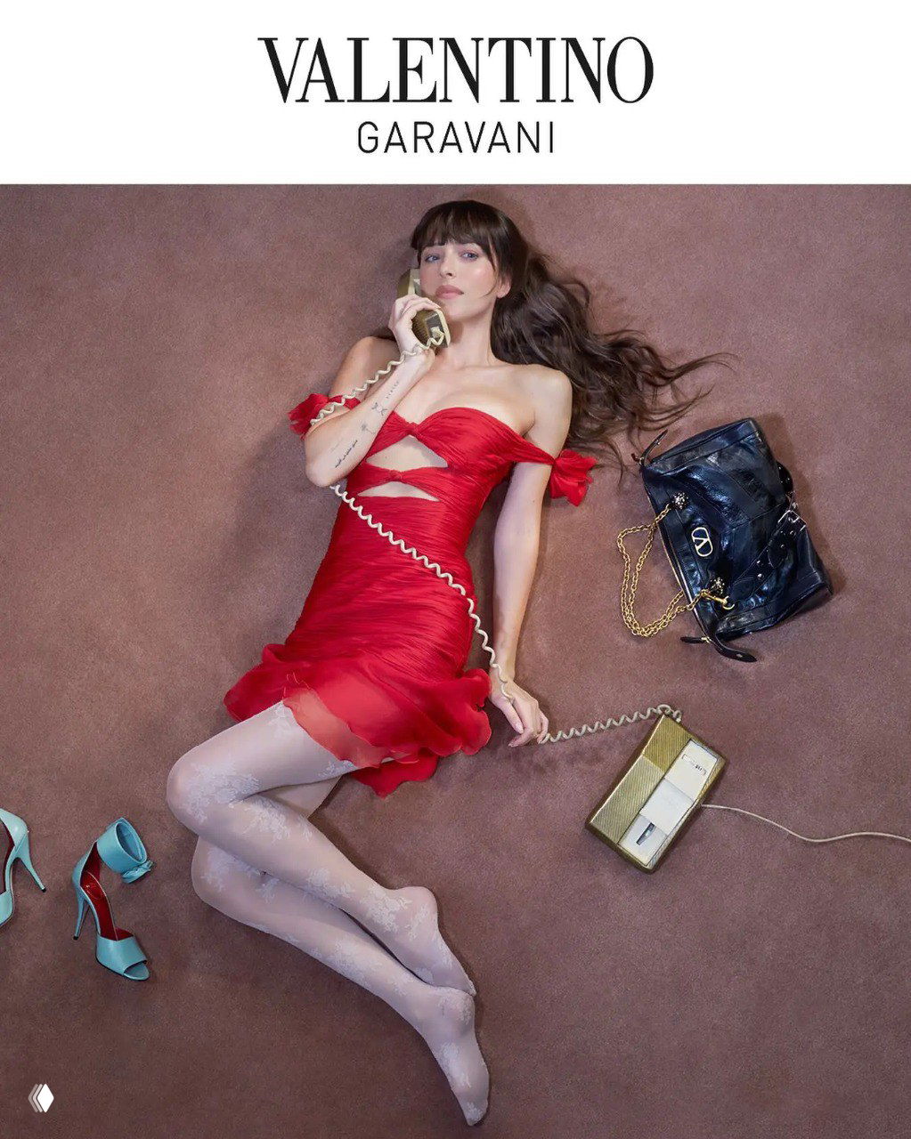 Дакота Джонсон лежит на ковре в красном платье Valentino, держит ретро-телефон; рядом сумка и туфли — кадр рекламной кампании.