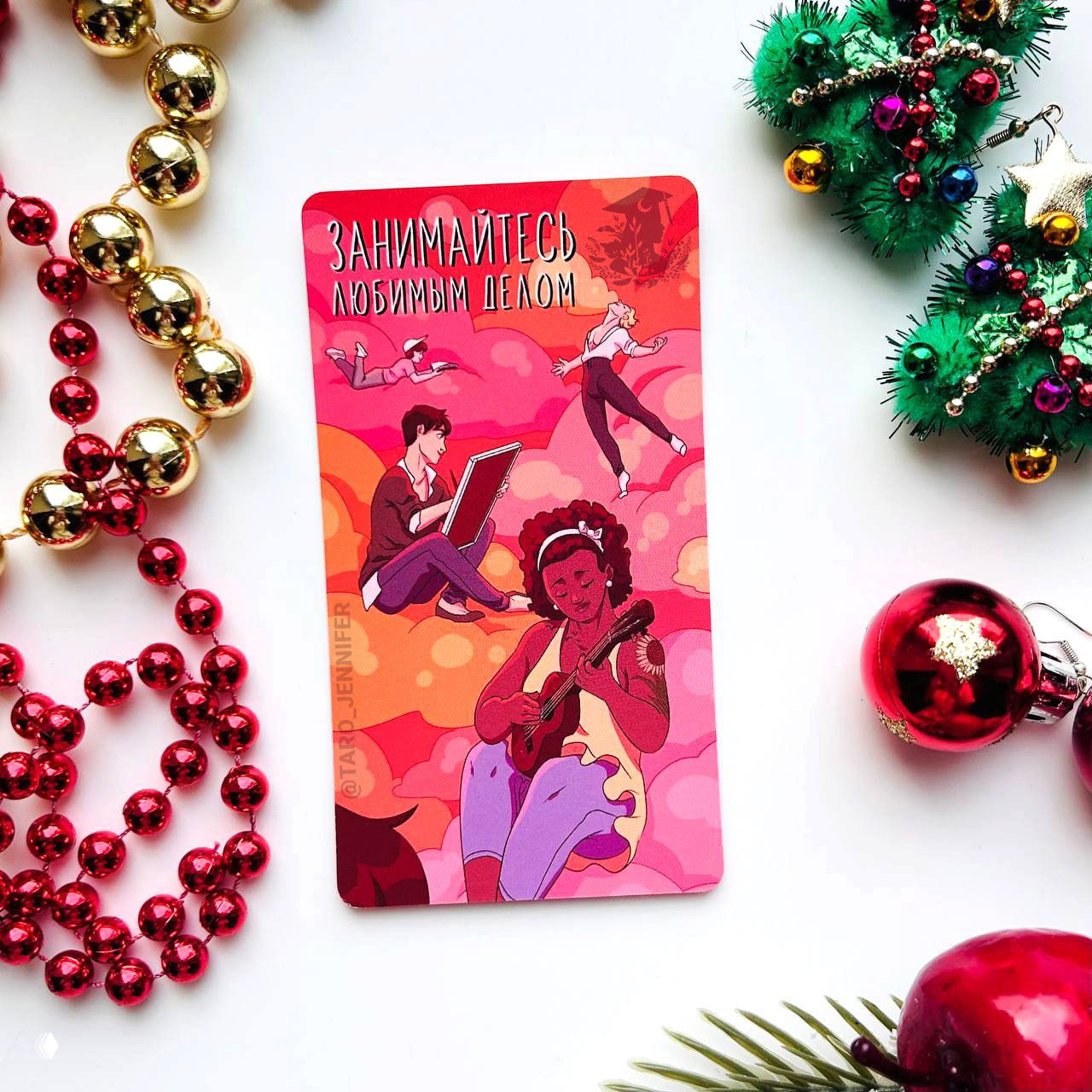 Карта Таро с надписью «Занимайтесь любимым делом» на белом фоне, рядом праздничные ёлочные игрушки и красные бусы.