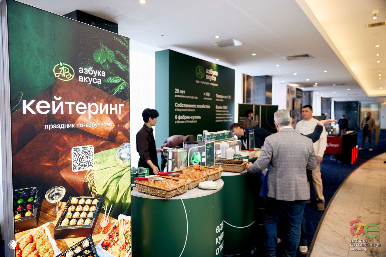 Стэнд кейтеринга «Азбука вкуса» с корзинами выпечки, угощениями и сотрудниками на стойке; участники подходят и выбирают закуски в коридоре площадки.