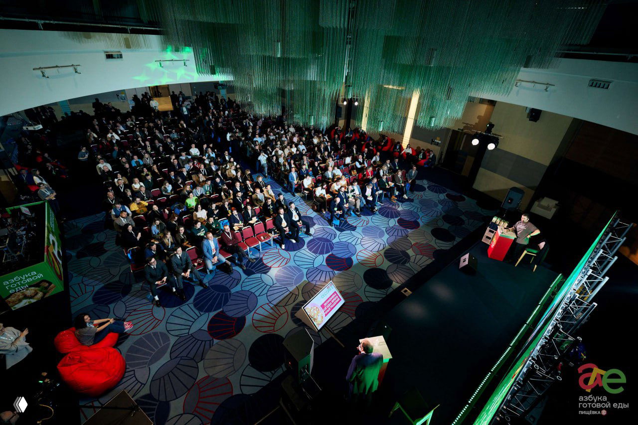 Общий вид конференц-зала сверху: заполнённый зал с рядами зрителей, сцена и экран внизу, большая аудитория слушает выступления форума.