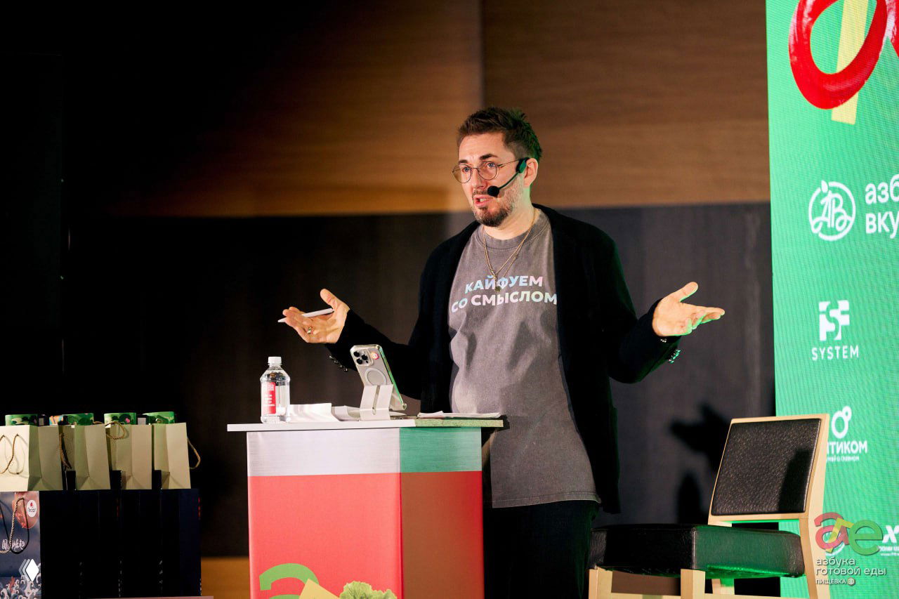 Крупный план спикера в гарнитуре на сцене: он жестикулирует во время выступления, на заднем плане видно брендирование и техническую сцену мероприятия.