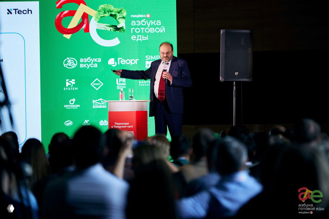 Докладчик у подиума на фоне зелёного фирменного экрана «азбука готовой еды», представляет слайды и обращается к слушателям в зале конференции.