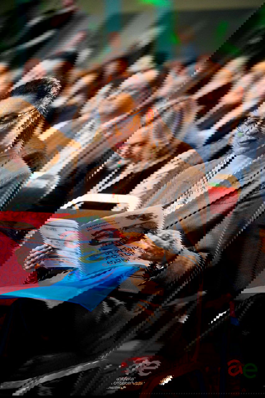 Участница в аудитории листает программу форума «Азбука готовой еды», вокруг видны другие слушатели; крупный план на буклет и бейдж участника.