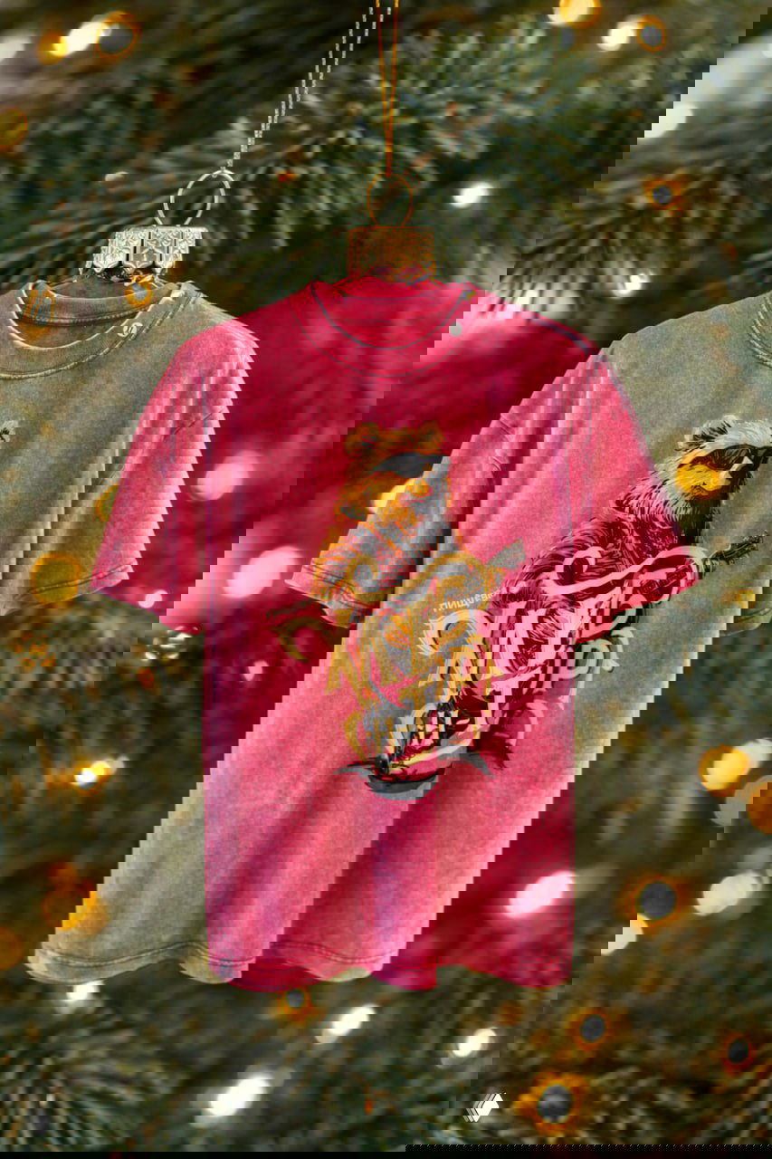 Игрушка‑футболка розового цвета с фирменным принтом мерча Пищёвки, подвешенная на ветке ели на фоне размытых гирлянд и огней.