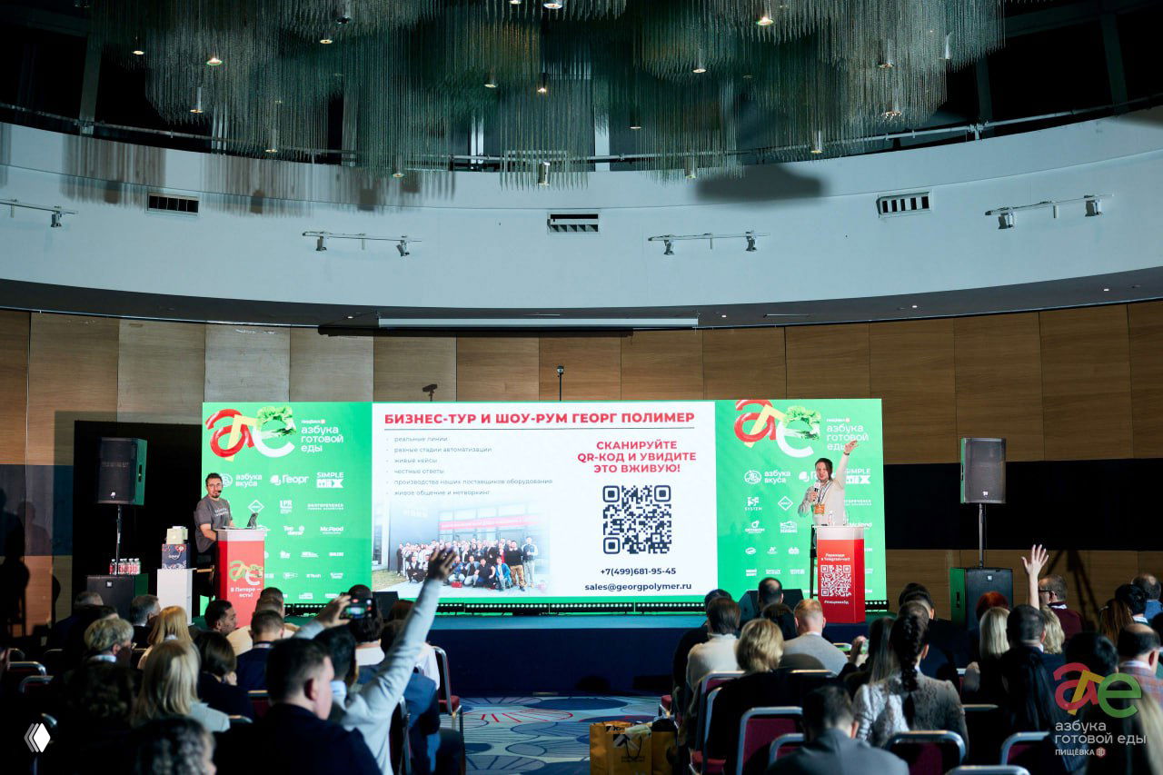 Широкий план сцены с презентацией и заметным QR‑кодом на слайде; зрители в зале, некоторые поднимают руки, видна сцена и аудитория.