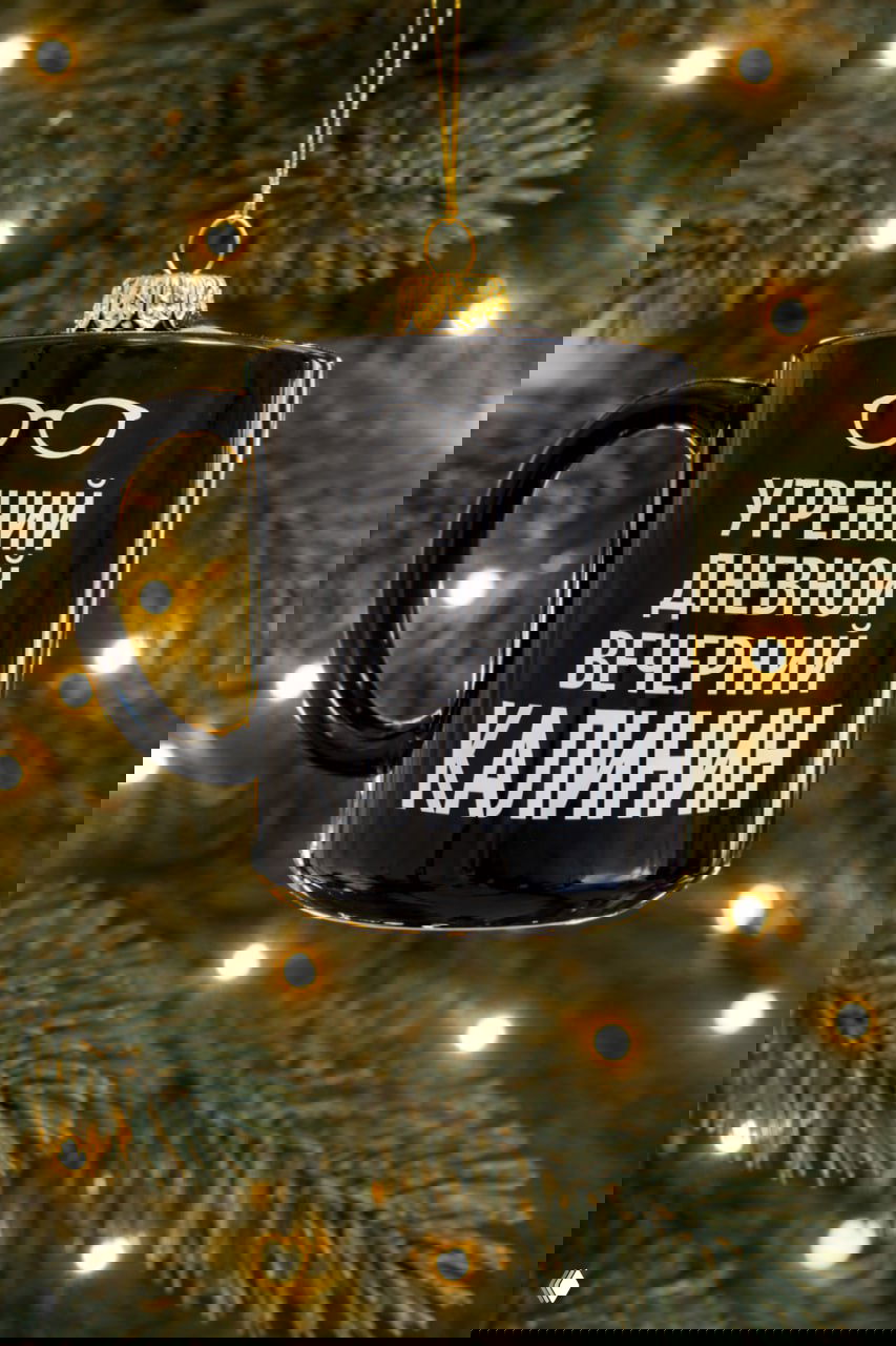 Чёрная кружка‑игрушка с текстом «УТРЕННИЙ ДНЕВНОЙ ВЕЧЕРНИЙ КАЛИНИН», подвешенная на еловой ветке — отсылка к спикеру Роману Калинину и коктейльной атмосфере.