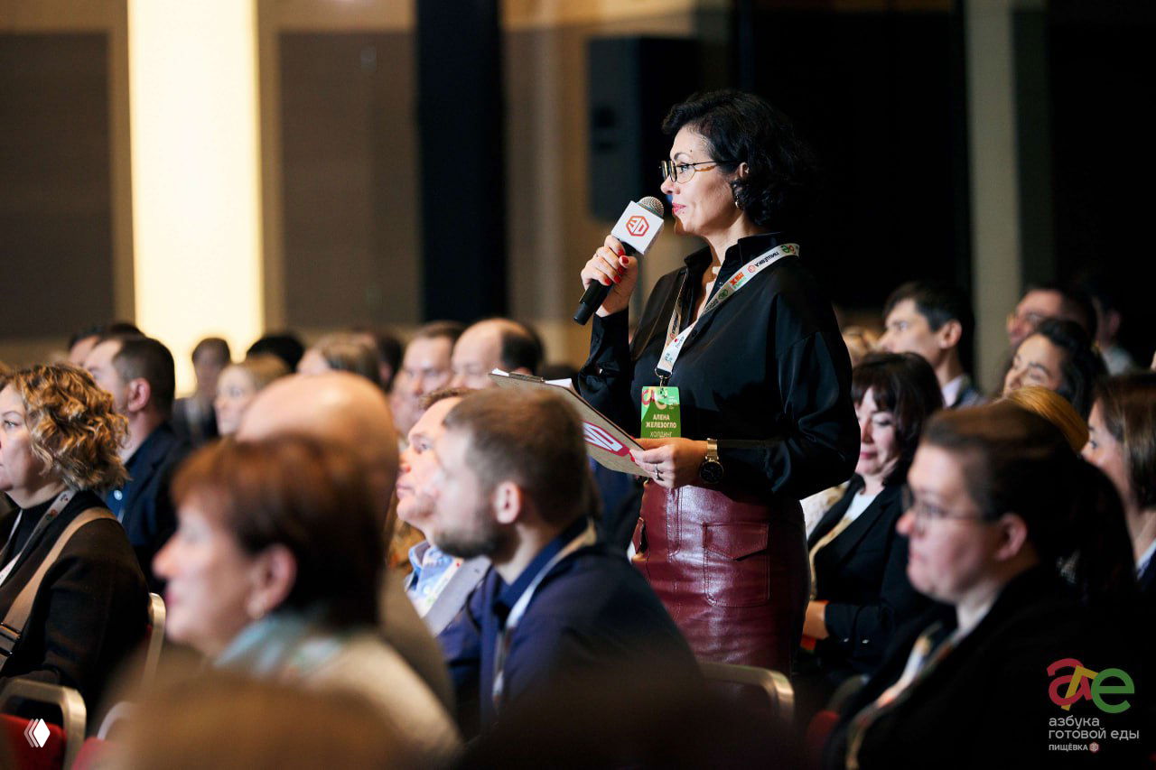 Женщина с микрофоном задаёт вопрос из зала: активное участие аудитории в сессии, вокруг сосредоточенные слушатели и деловая атмосфера конференции.