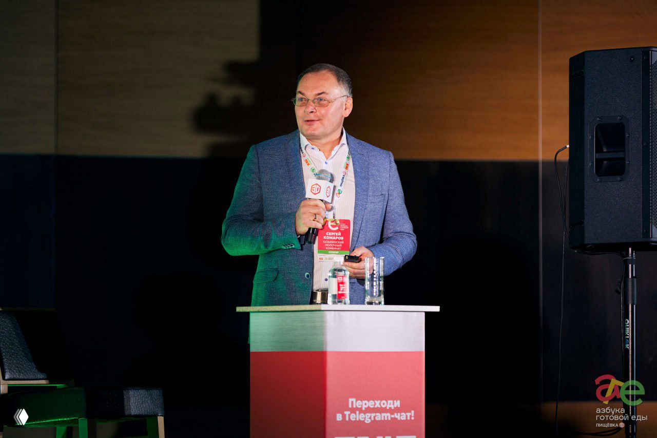 Спикер в деловом костюме у подиума выступает перед аудиторией, видна брендированная зелёная панель и реквизит на сцене мероприятия.