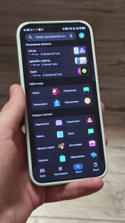 Смартфон под углом в светлом чехле; на экране тёмная тема и набор иконок приложений, фон — пол и бытовая обстановка, подчёркивающие реальные условия использования.