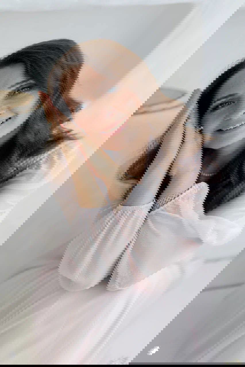 Портрет улыбающейся Марии Бражниковой в светлом наряде, руки сложены у лица, мягкий интерьерный фон, дружелюбный выразительный взгляд.