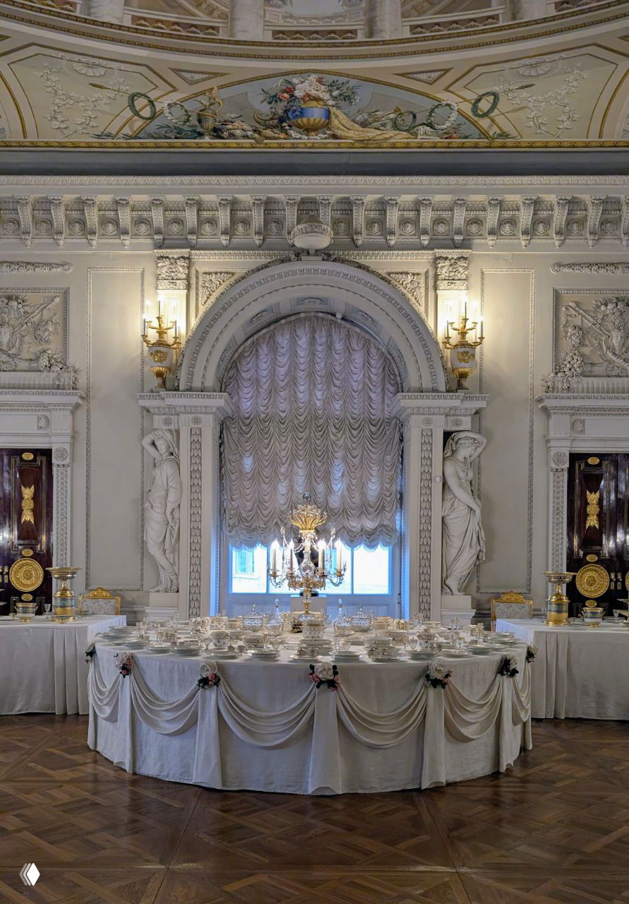 Большой парадный зал с сервировкой: овальный стол, фарфоровая посуда и хрустальные люстры на фоне лепнины и высоких окон дворца.