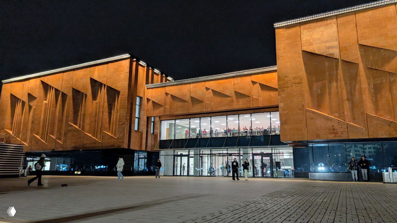 Входная часть библиотеки с просторной площадью и стеклянными секциями в основании здания; люди заходят и выходят из холла вечером.