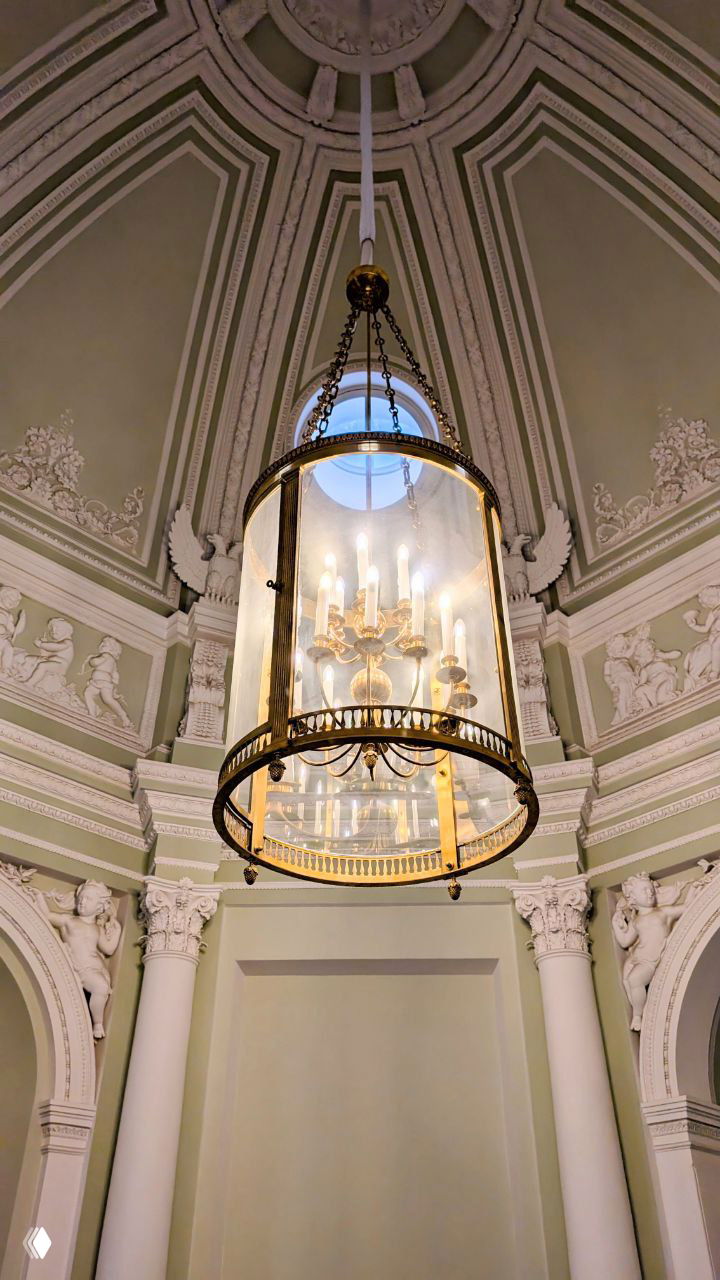 Крупный план стеклянной фонарной люстры-«барабана» в куполе Большого Гатчинского дворца: светильник, цепи и декоративная лепнина вокруг.