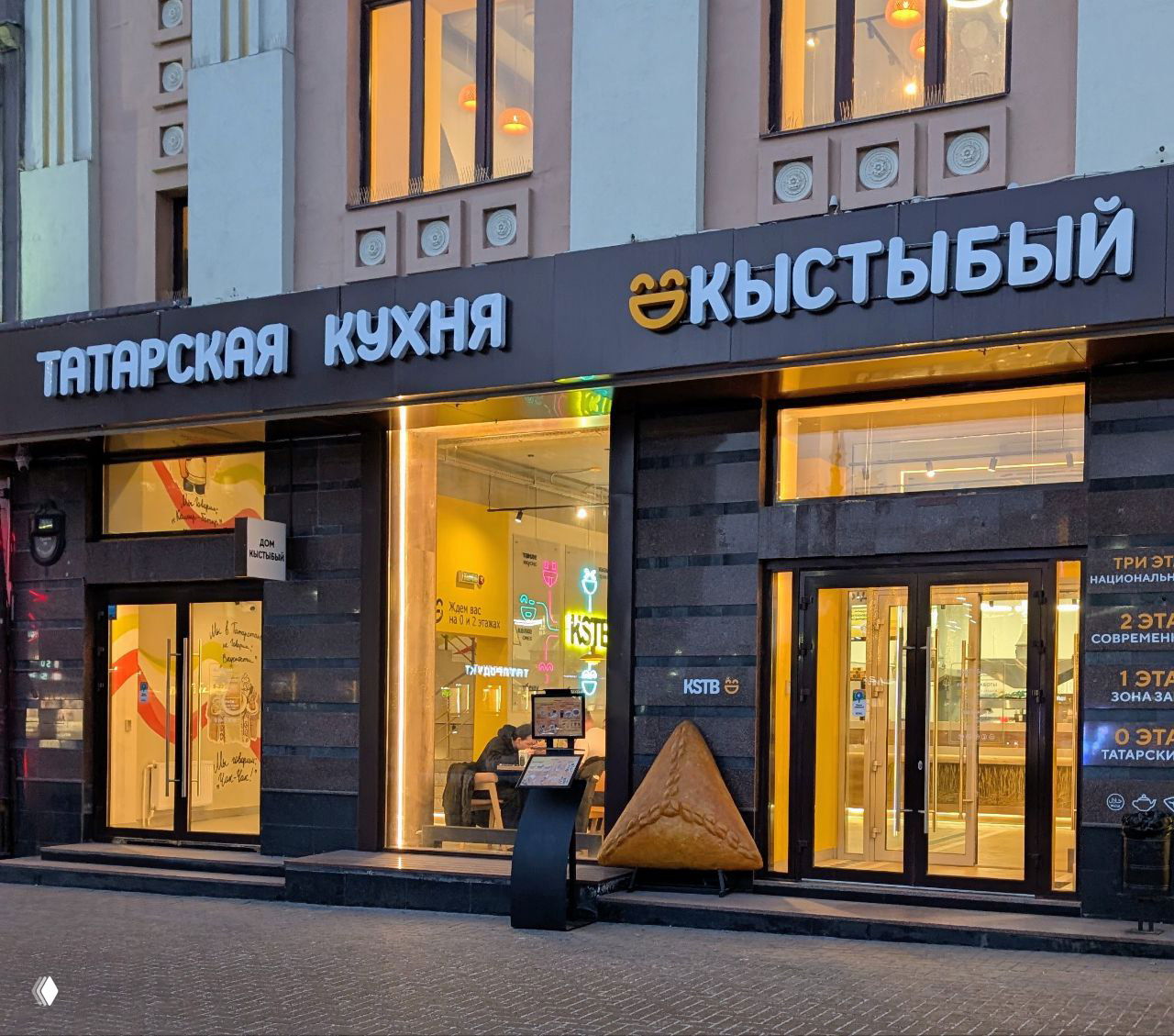 Вечерний фасад заведения «Кыстыбый» с надписью 'Татарская кухня' и большой декорацией в виде треугольного эчпочмака у входа, тёплая подсветка и витрины.