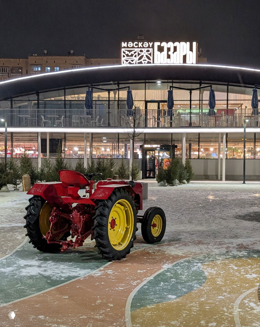 Ночной кадр площади перед Московским рынком: светящаяся вывеска могучего павильона 'Московские базары' и красный декоративный трактор на заснеженной площадке в переднем плане.