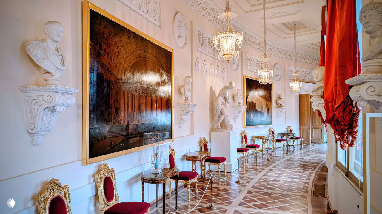 Парадная галерея с бюстами, картинами в золочёных рамах, рядами кресел и декоративными люстрами вдоль светлого коридора.