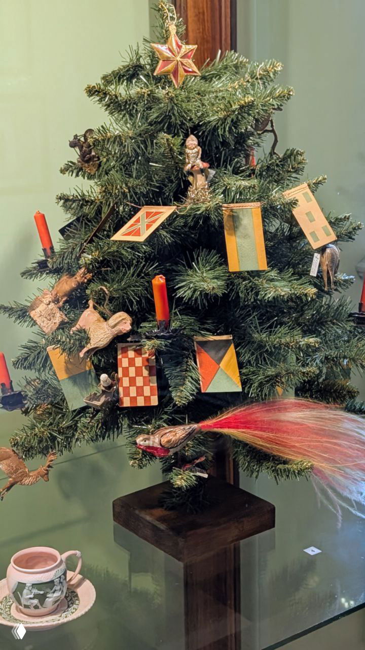 Небольшая музейная рождественская ёлка с традиционными деревянными игрушками, красными свечами и чашкой чая на столе рядом — атмосферная деталь интерьера.