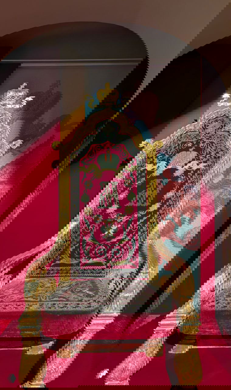 Тронное кресло на красном подиуме с вышивкой вензеля и короной, фронтальный портретный ракурс экспоната в Троицкой церкви Гатчинского дворца