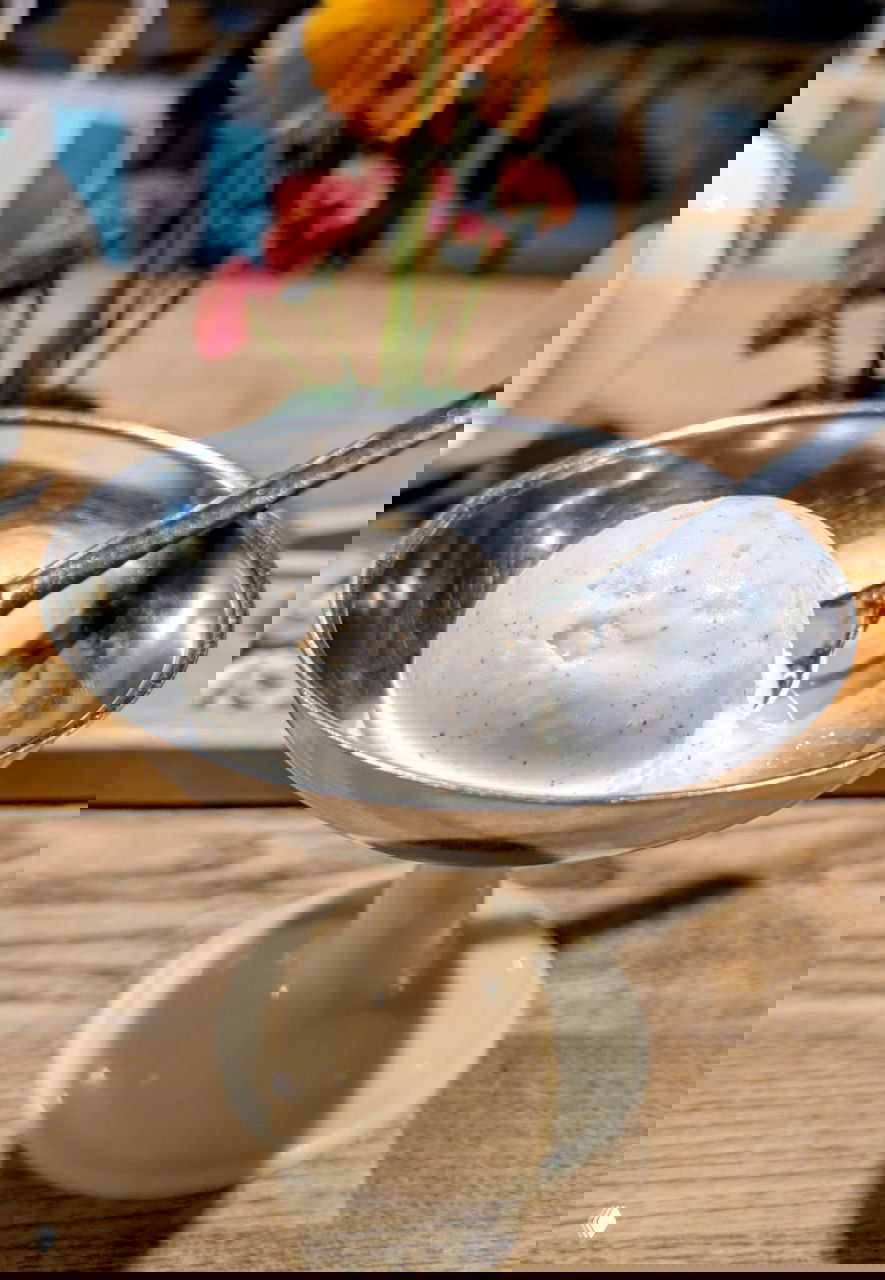 Шарик белого мороженого в металлической креманке на деревянном столе; фоном — чайная посуда и цветы, отражающие ресторанную атмосферу.