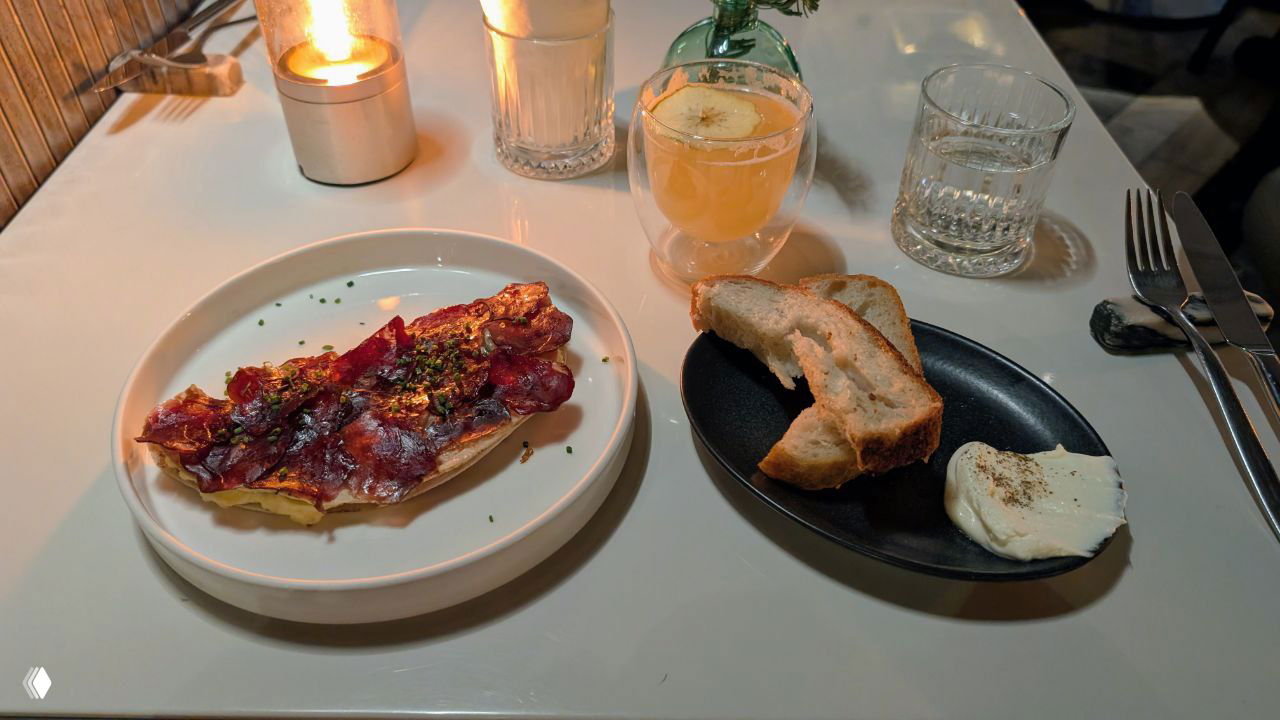 Брускетта с тонкими ломтиками вяленой конской колбасы, тарелка с хлебом и маслом, тёплая ленивая ресторанная сцена в мягком свете.