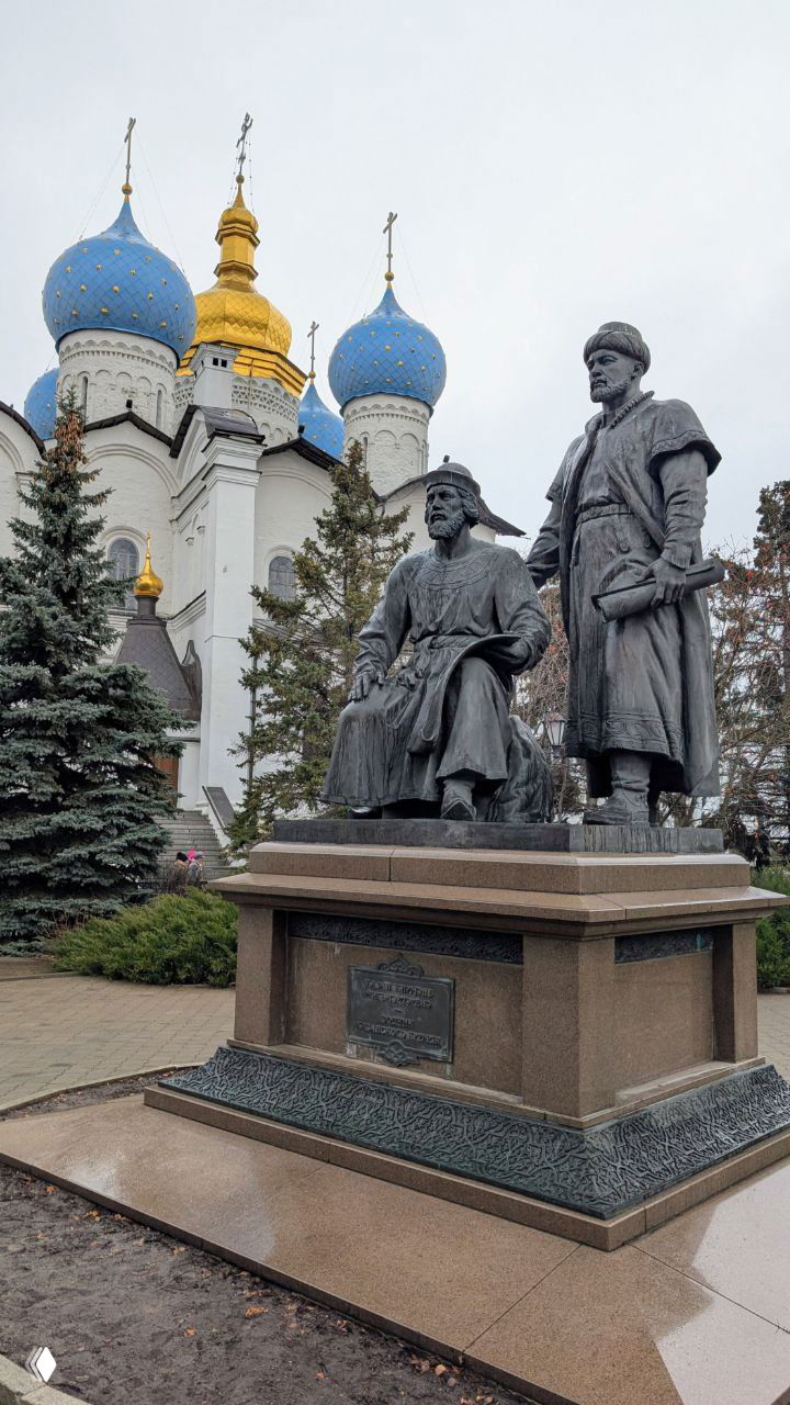 Памятник и купола Благовещенского собора в Казани: бронзовая скульптура на переднем плане и голубые луковки с золотыми крестами позади.