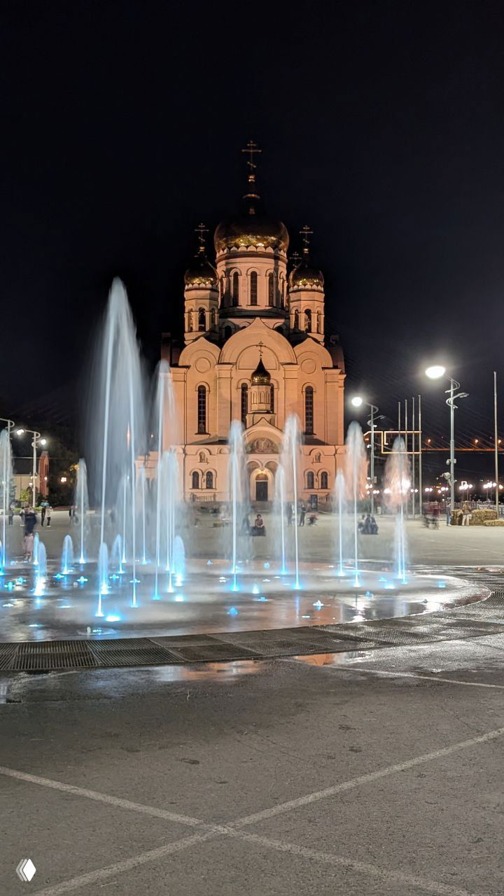 Ночной вид на фонтан и освещённый собор во Владивостоке: голубые струи фонтана на переднем плане и золотые купола храма на заднем