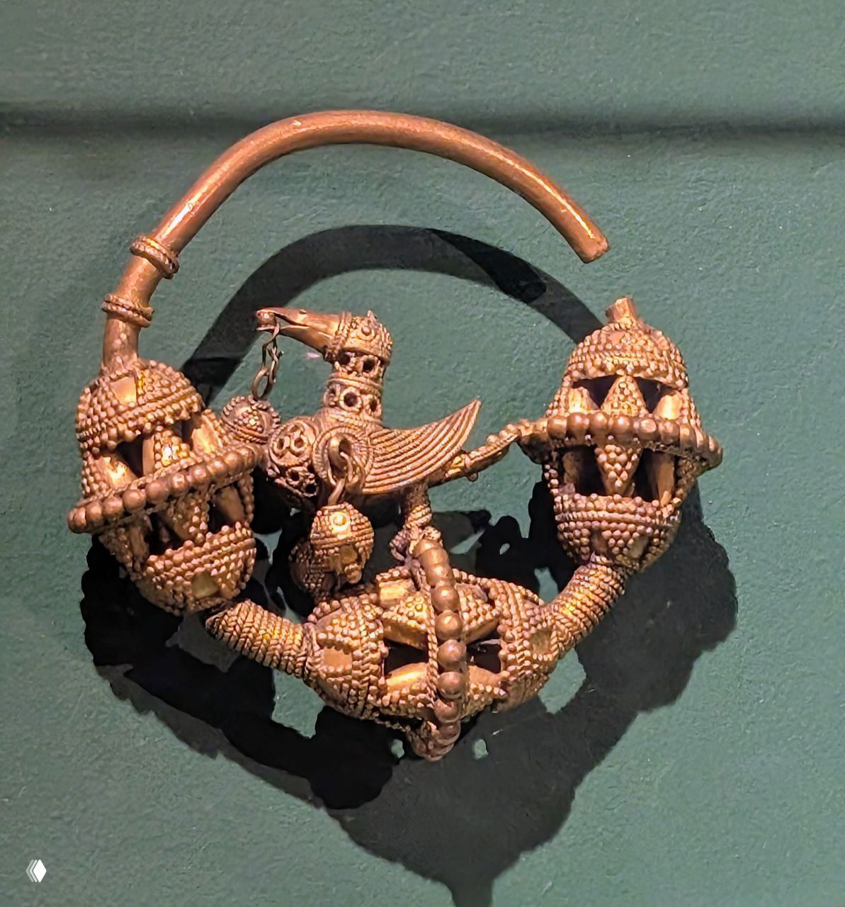 Малый музейный экспонат — древняя ювелирная находка с изображением утки и сложной филиграни, выставленная на зелёном фоне музейной витрины.