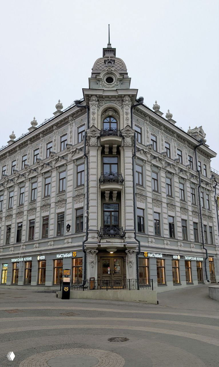 Угловой фасад исторического многоэтажного дома с декоративными элементами и балконами на улице Баумана, видны витрины магазинов на первом этаже.